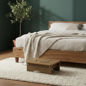 May include: A wooden bed frame with a light-colored blanket and pillows. A small wooden step stool with a cutout is in front of the bed. The room has a green wall and a white rug.