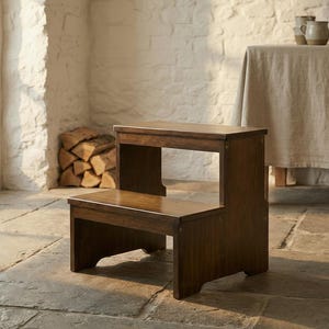 May include: A two-step wooden step stool with a rich, dark brown finish. The stool has a rectangular top step and a lower step, connected by a side panel. The design is simple and classic, suitable for reaching higher shelves or as a decorative accent.