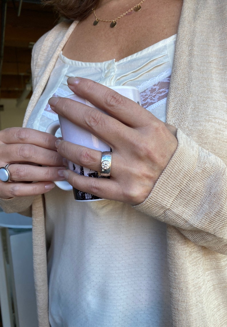 May include: A silver ring with a large, round, clear stone set in a textured band. The ring is worn on the index finger of a person's hand.