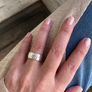 May include: A silver and gold ring with a large, clear gemstone set in the centre. The ring has a hammered texture and a wide band.