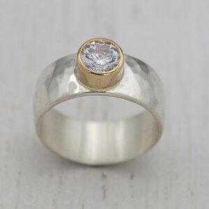 May include: A silver and gold ring with a hammered texture and a clear round gemstone.
