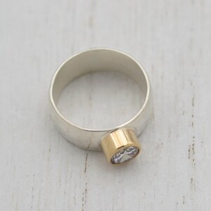 May include: A silver ring with a gold band and a clear gemstone. The ring is set on a white wooden surface.