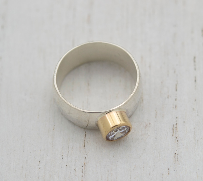 May include: A silver ring with a gold band and a clear gemstone. The ring is set on a white wooden surface.