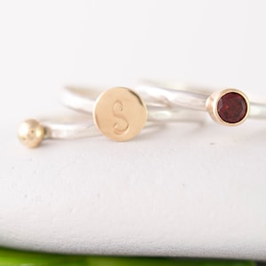 May include: Three silver rings with gold accents. One ring has a small gold ball, one has a round gold disc with the letter "S" engraved on it, and one has a small red gemstone.