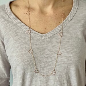 May include: A long gold chain necklace with multiple open heart charms. The necklace is worn by a person with a light grey shirt.