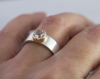 May include: A silver and gold ring with a clear gemstone set in a gold bezel. The ring is worn on a person's finger.
