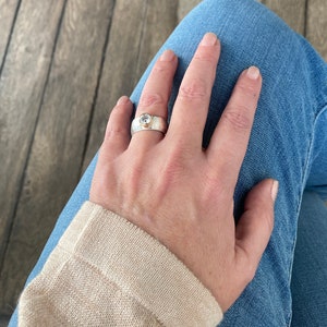 May include: A silver ring with a round, clear gemstone set in a gold bezel. The ring is worn on a person's finger.
