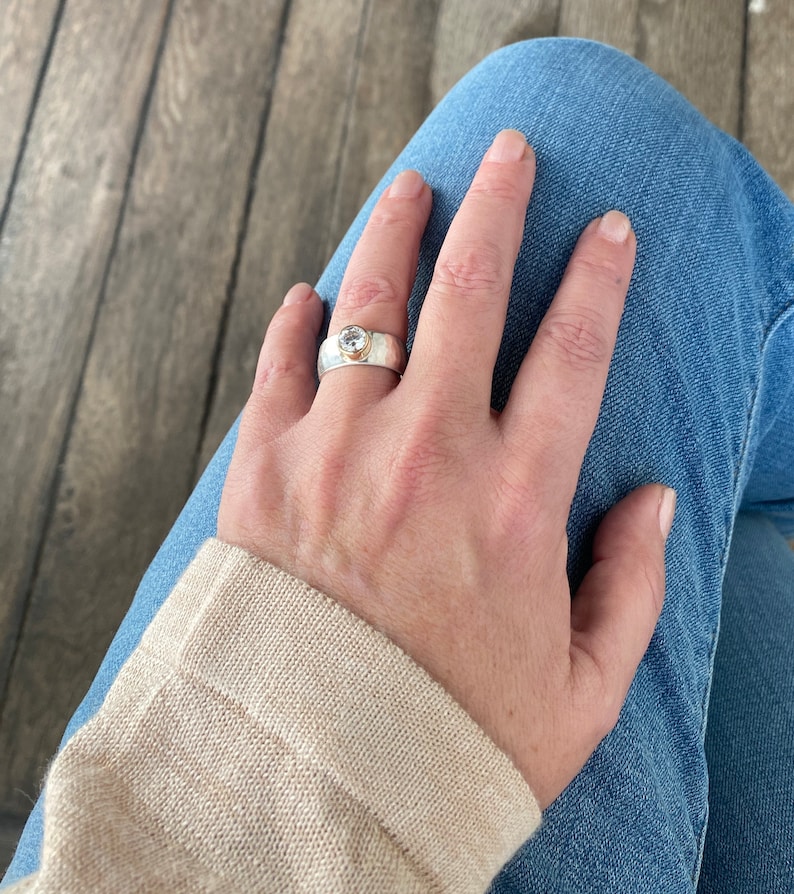 May include: A silver ring with a round, clear gemstone set in a gold bezel. The ring is worn on a person's finger.