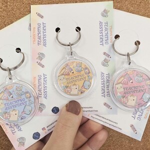 Può includere: Tre portachiavi circolari in acrilico trasparente con anelli in metallo, ciascuno fissato su un cartoncino. Gli inserti presentano disegni di materiale scolastico e il testo Thank You Teaching Assistant in un design dai toni pastello.
