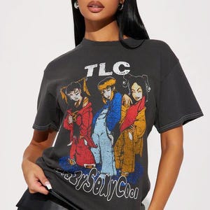 May include: Dark gray t-shirt featuring a graphic of the music group TLC. The graphic shows three cartoon figures in colorful outfits. The text "TLC" is above the graphic, and "CrazySexyCool" is below.