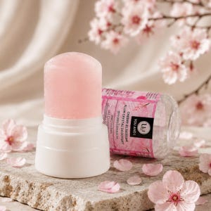 Könnte beinhalten: Ein zylindrischer, hellrosa Kristall-Deodorantstift mit weißem Kunststoffsockel. Das Etikett zeigt den Text "YOU AND I" und "CHERRY BLOSSOM ALUM CRYSTAL DEODORANT" mit floralen Grafiken. Das Produkt befindet sich in einem durchsichtigen Behälter.