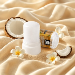 Könnte beinhalten: Ein weißer, zylindrischer Kristall-Deodorantstift mit einem Etikett, auf dem You and I Coconut Alum Crystal Deodorant steht. Der Behälter ist von einer halbierten braunen Kokosnuss mit weißem Fruchtfleisch und drei weißen Plumeria-Blüten umgeben.