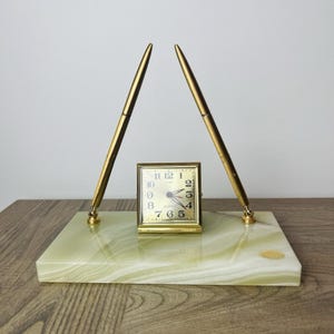 May include: A desk set featuring a rectangular light green onyx base with two gold-toned pens held in upright swivel mounts. A square gold-toned analog clock with a white face and black numerals is positioned in the center of the stone base.
