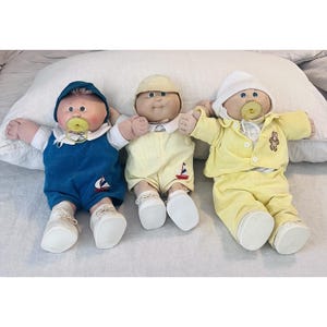 Pode incluir: Três bonecos com rostos redondos e cabelo de lã. O boneco da esquerda veste um macacão azul com um veleiro e um gorro azul. O do meio veste jardineiras amarelas com um veleiro e um gorro amarelo. O da direita veste um pijama amarelo e gorro branco.