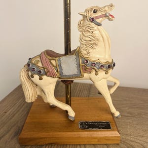 May include: A cream-colored carousel horse figurine with a pink saddle, purple gemstone accents, and a gold-tone pole, mounted on a rectangular wooden base with a metal plaque reading P.J.'s Carousel Collection Replica of Early Illion.