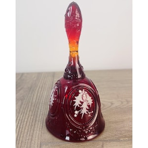 Puede incluir: Una campana de cristal rojo intenso con un mango alto y delgado con detalles florales en relieve. El cuerpo principal de la campana está decorado con paneles circulares que contienen diseños grabados en blanco de hojas y bayas de acebo.