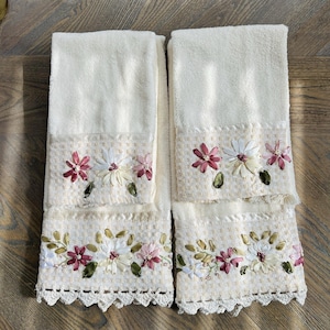 May include: Two cream-colored hand towels with floral embroidery. The towels feature a decorative band with embroidered flowers in shades of pink, white, and green. The bottom edge has a delicate crocheted lace trim, adding a touch of elegance.