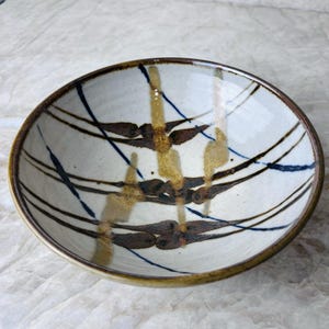 May include: A round ceramic bowl with a speckled off-white glaze featuring hand-painted abstract patterns. The design includes dark brown horizontal leaf-like shapes, vertical golden-brown brushstrokes, and thin dark blue diagonal lines.