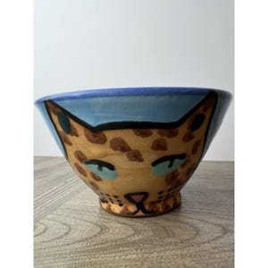 Puede incluir: Un cuenco de cerámica pintado a mano con un diseño de leopardo. El cuenco tiene un borde azul claro y una base beige con manchas marrones. La cara del leopardo está delineada en negro, con ojos azules y una nariz rosa. El cuenco está sobre una superficie de madera.
