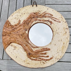 May include: Circular ceramic mirror featuring a cream backdrop and a brown, tree-like design. The mirror has a circular shape with a brown frame and a leather hanging loop. The ceramic has a speckled appearance.