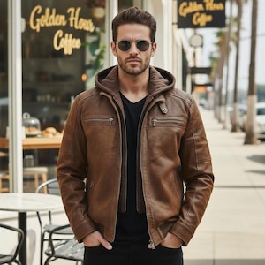 May include: A brown leather jacket with a hood and multiple zippered pockets. The jacket is worn over a black shirt. The person in the image is wearing sunglasses. The background includes a coffee shop and a sidewalk.