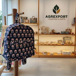 以下が含まれることがあります： ピンクの花柄のネイビーブルーのバックパックが木製のディスプレイに吊るされています。店内には、バッグや装飾品など、さまざまな手作りのアイテムが並んだ棚があります。「AGREXPORT - HANDLOOM & CRAFTS」というお店のロゴが見えます。