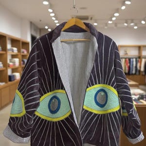 May include: A dark navy blue jacket with a unique eye design. The jacket features large embroidered eyes in shades of blue, yellow, and green. The interior lining is striped, and the jacket is hanging on a wooden hanger.