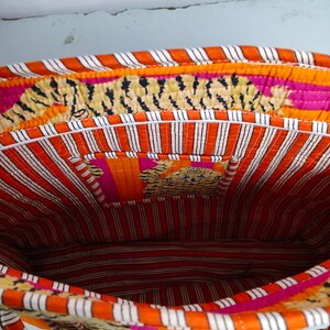 May include: An open, quilted tote bag with a vibrant interior. The bag features a striped orange and white lining, with a small pocket. The exterior trim has a tiger print design, with pink and orange accents. The bag's handles are also visible.