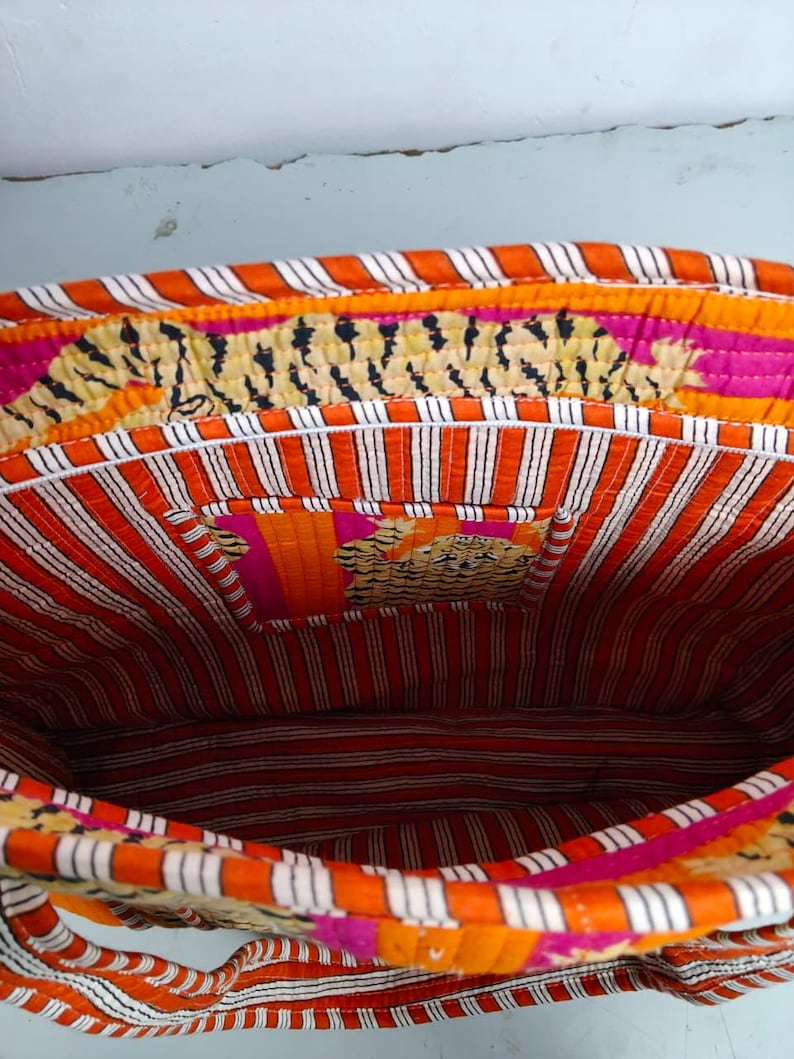 May include: An open, quilted tote bag with a vibrant interior. The bag features a striped orange and white lining, with a small pocket. The exterior trim has a tiger print design, with pink and orange accents. The bag's handles are also visible.