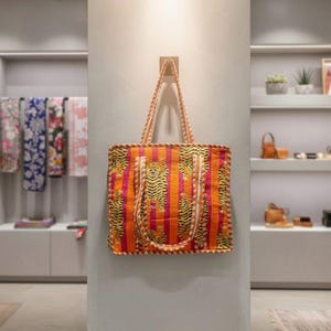 May include: An orange and pink tote bag with a striped pattern and tiger print accents. The bag has long handles and is hanging on a wall. The bag is part of a collection of bags and accessories.