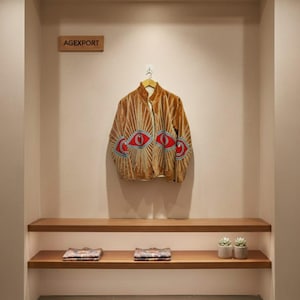 Op de afbeelding: Een bruin fluwelen jasje met een uniek oogontwerp, tentoongesteld op een houten hanger. De jas heeft rode en blauwe geborduurde ogen en zonnestralenpatronen. Het bord boven de jas luidt "AGEXPORT".