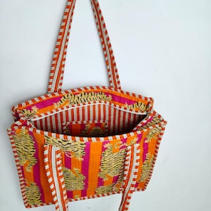 May include: A vibrant tote bag with a tiger print and striped accents. The bag features orange, pink, and white colors. The handles are striped in orange and white. The bag is open, revealing the interior lining.