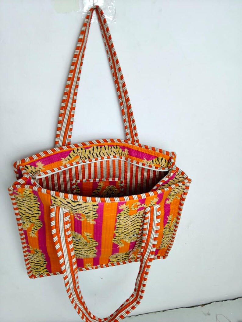 May include: A vibrant tote bag with a tiger print and striped accents. The bag features orange, pink, and white colors. The handles are striped in orange and white. The bag is open, revealing the interior lining.