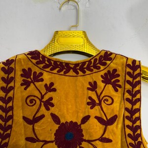 May include: Mustard yellow vest with intricate maroon floral embroidery. The vest features a round neckline and a sleeveless design. The embroidery includes a central flower with a blue center and detailed leaf patterns.