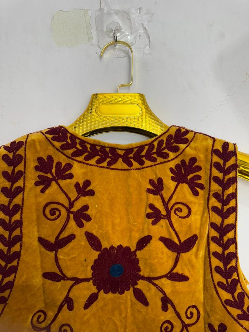 May include: Mustard yellow vest with intricate maroon floral embroidery. The vest features a round neckline and a sleeveless design. The embroidery includes a central flower with a blue center and detailed leaf patterns.