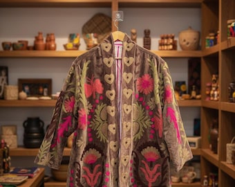 Pode incluir: Casaco de veludo castanho com bordados florais intrincados em rosa, verde e dourado. O casaco apresenta um design em forma de coração na frente e nas mangas, criando uma estética boêmia. O casaco está pendurado num cabide de madeira.