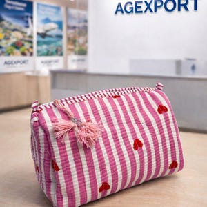 Op de afbeelding: Een roze en witte gestreepte rechthoekige cosmetische tas met kleine rode hartaccenten. De tas heeft een roze kwast en ligt op een licht houten oppervlak. De achtergrond bevat de tekst "AGEXPORT".