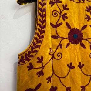 May include: Mustard yellow vest with intricate maroon floral embroidery. The sleeveless garment features a round neckline and is displayed on a gold hanger. The embroidery includes floral and vine patterns.