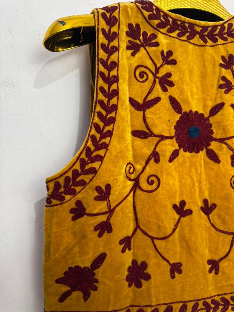 May include: Mustard yellow vest with intricate maroon floral embroidery. The sleeveless garment features a round neckline and is displayed on a gold hanger. The embroidery includes floral and vine patterns.