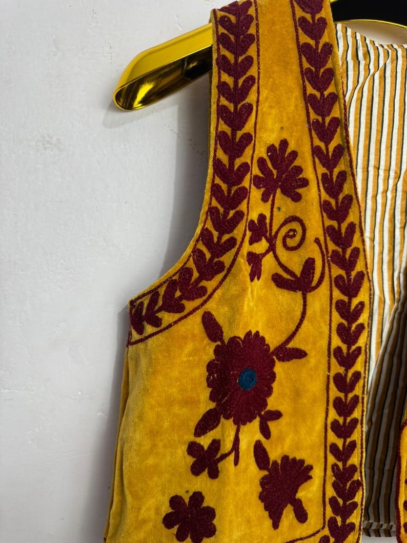May include: Mustard yellow vest with burgundy floral embroidery and leaf trim. The vest has a striped lining and is hanging on a gold hanger. The embroidery features a large flower with a teal center.