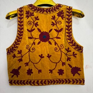 May include: Mustard yellow vest with burgundy floral embroidery. The vest features a central flower design with a teal center and intricate vine and leaf patterns. The edges are trimmed with a burgundy leaf pattern.