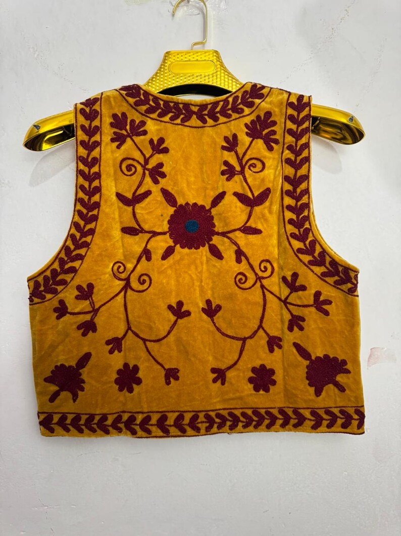 May include: Mustard yellow vest with burgundy floral embroidery. The vest features a central flower design with a teal center and intricate vine and leaf patterns. The edges are trimmed with a burgundy leaf pattern.