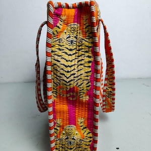 May include: A quilted tote bag with a vibrant tiger print in shades of orange, pink, and yellow. The bag features striped handles and trim in red, white, and orange. The bag is rectangular and has a structured shape.