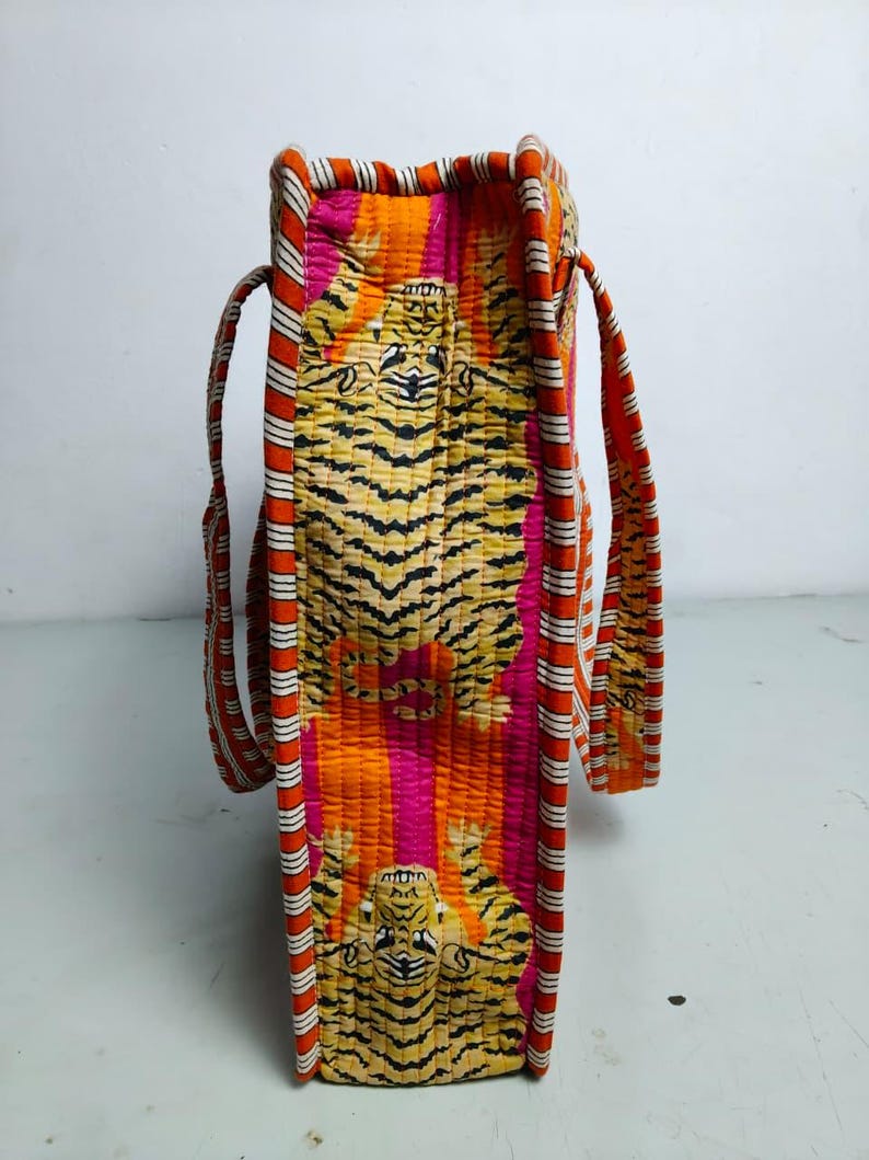 May include: A quilted tote bag with a vibrant tiger print in shades of orange, pink, and yellow. The bag features striped handles and trim in red, white, and orange. The bag is rectangular and has a structured shape.