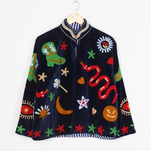 Pode incluir: Uma jaqueta de veludo azul-marinho escuro com motivos bordados coloridos, incluindo uma bota verde, uma cobra vermelha, uma lua crescente amarela, um sol, estrelas, corações e olhos. A jaqueta tem gola mandarim e forro listrado.