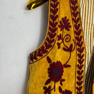 May include: Mustard yellow vest with burgundy floral embroidery and leaf trim. The vest has a striped lining and is hanging on a gold hanger. The embroidery features a large flower with a teal center.