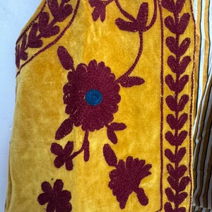 May include: Mustard yellow vest with burgundy floral embroidery and a striped fabric backing. The vest features a velvet texture and intricate embroidered floral designs, with a blue center in the largest flower.
