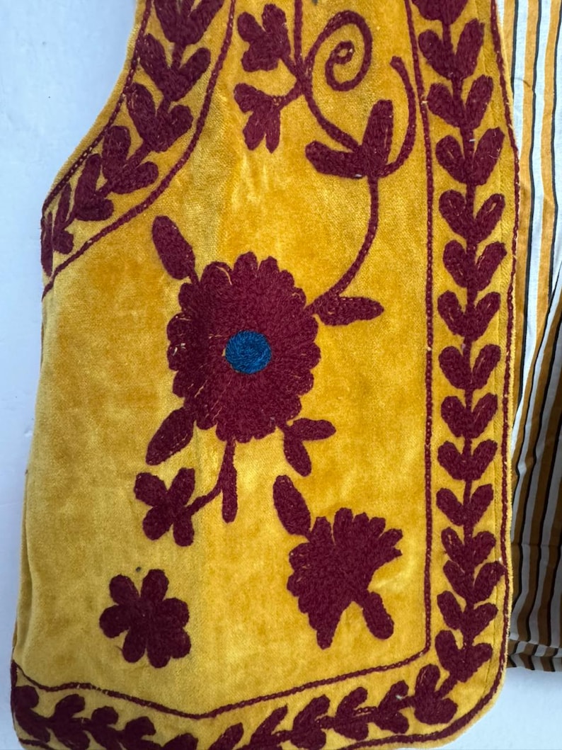 May include: Mustard yellow vest with burgundy floral embroidery and a striped fabric backing. The vest features a velvet texture and intricate embroidered floral designs, with a blue center in the largest flower.