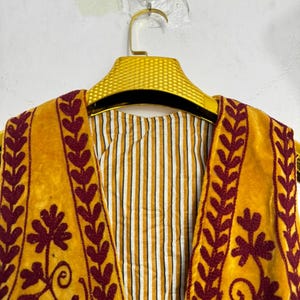 May include: Close-up of a mustard yellow vest with burgundy floral embroidery and leaf patterns. The vest is hanging on a gold hanger, revealing a striped lining in white and yellow. The garment is made of a soft, velvety material.