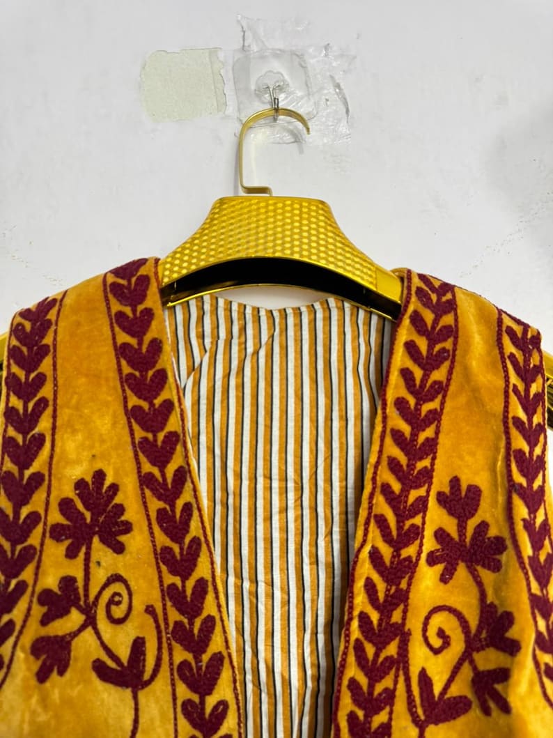 May include: Close-up of a mustard yellow vest with burgundy floral embroidery and leaf patterns. The vest is hanging on a gold hanger, revealing a striped lining in white and yellow. The garment is made of a soft, velvety material.